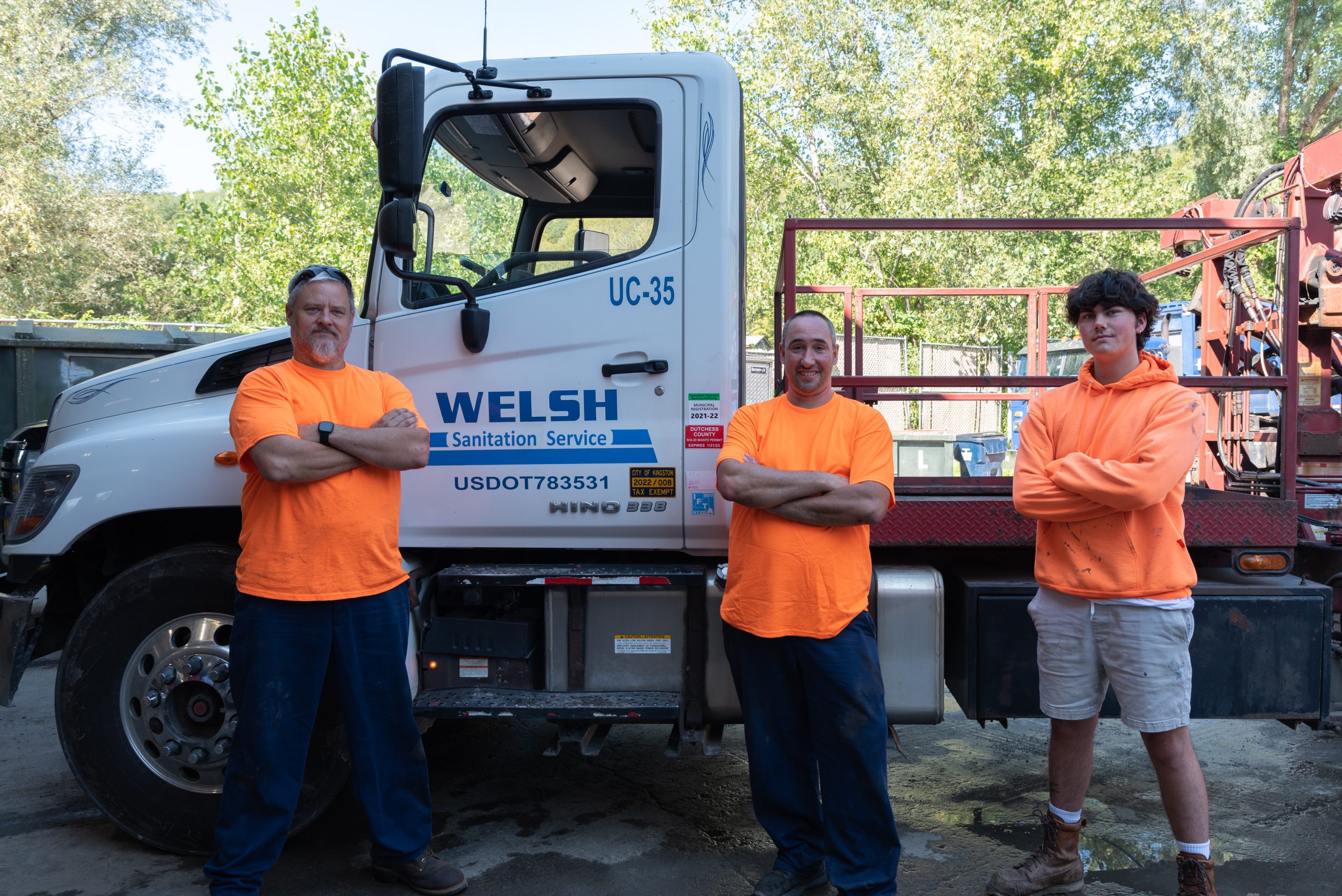 Three men in orange shirts standing in front of Welsh Sanitation truck