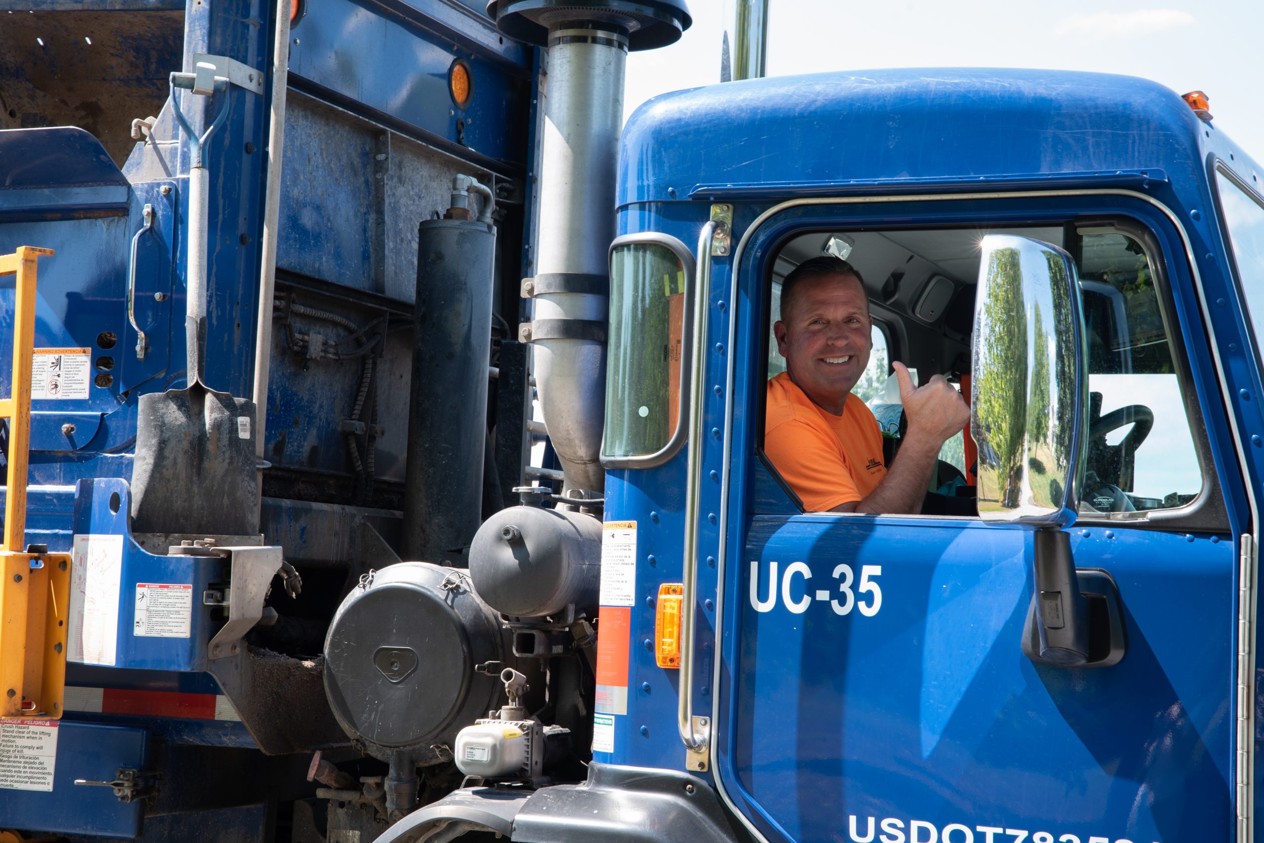 Male employee sitting in blue sanitation truck while smiling and giving thumbs up